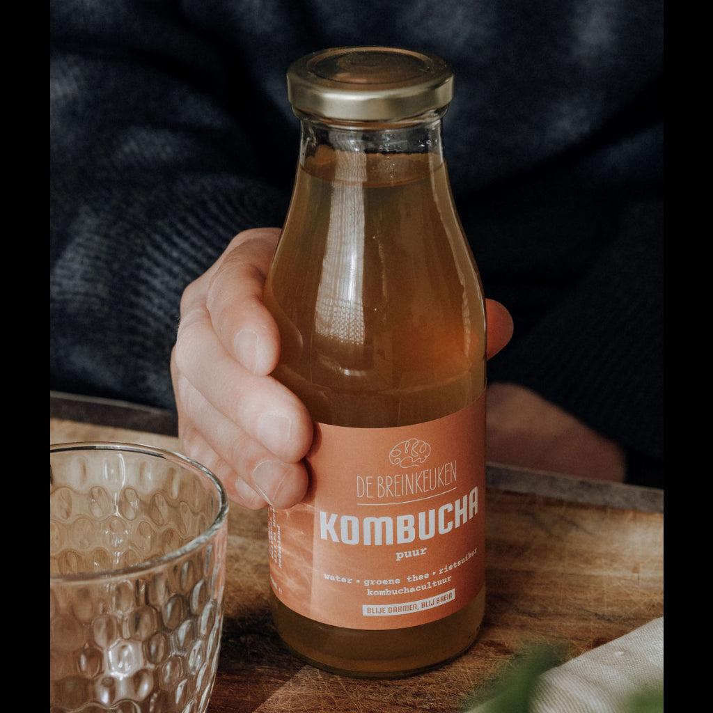 Fles pure kombucha van De Breinkeuken vastgehouden in een hand naast een leeg glas